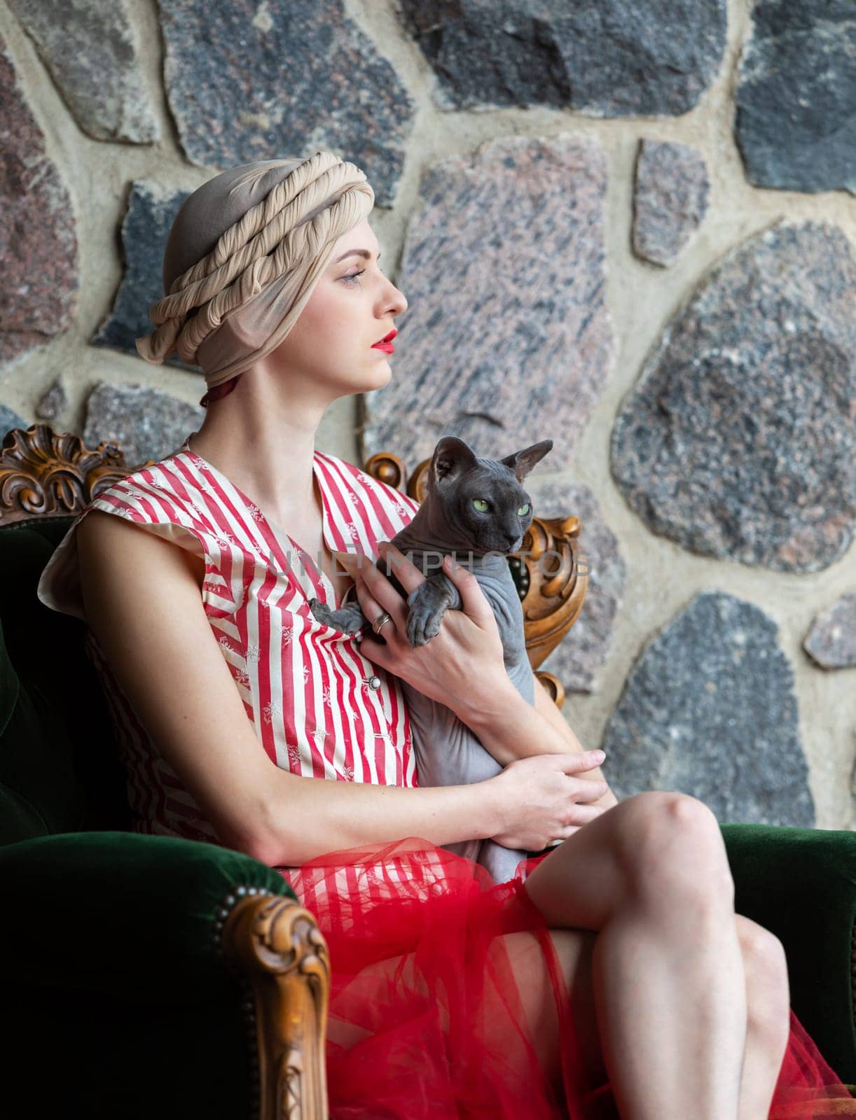 A woman in a fashionable dress and head wrap sits gracefully on an antique chair. She holds a cat closely, posing in front of a textured stone wall, creating an elegant atmosphere.
