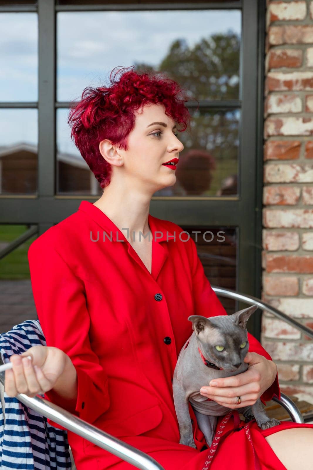 A woman with vibrant red hair wears a striking red outfit while sitting comfortably and holding a gray cat. The background features a bright, modern interior with large windows.