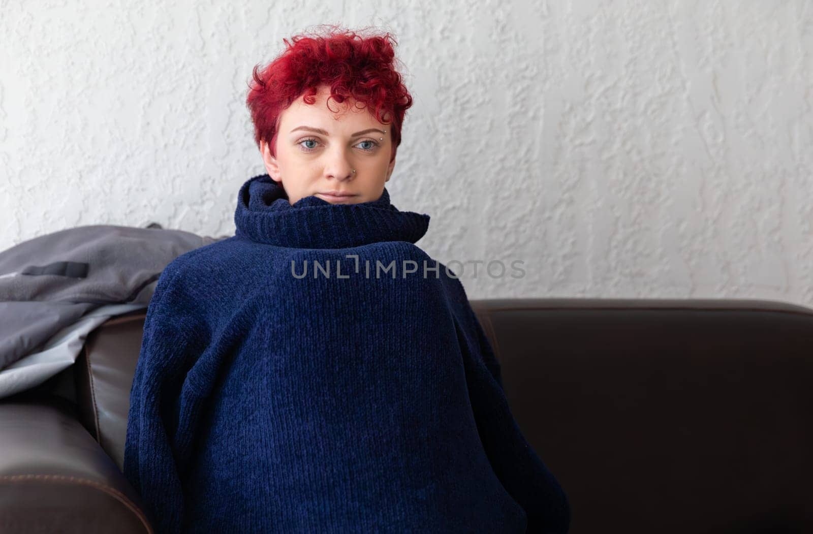 A person with curly red hair sits comfortably on a dark brown couch, wrapped in a large navy sweater. The light and textured wall adds a cozy feel to the indoor environment.
