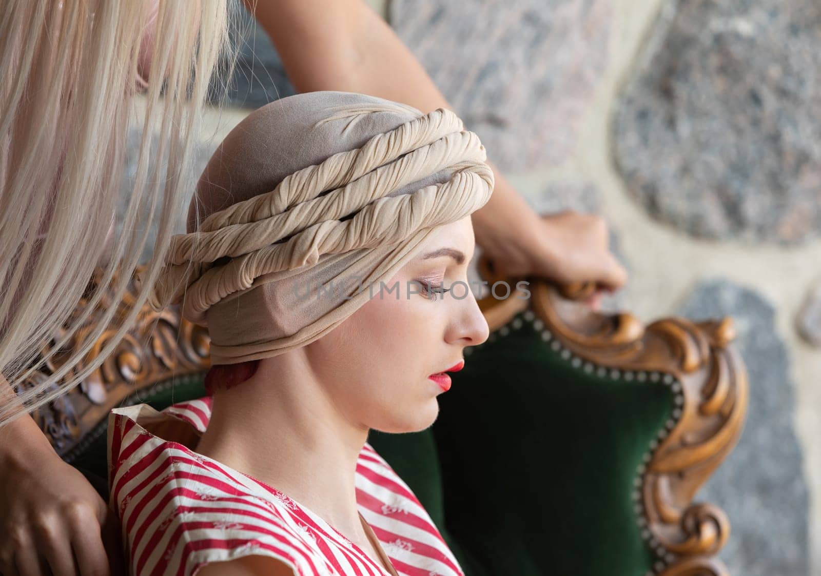 A woman with a refined look sits gracefully on a vintage chair. A hairstylist carefully works on her hair, creating an intricate updo.