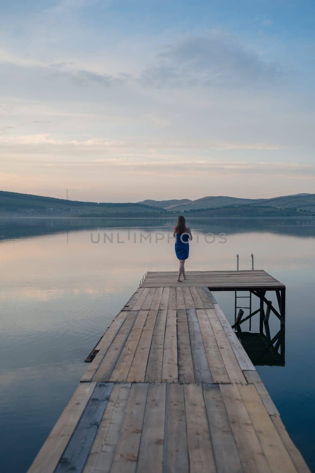 A young woman with wet hair, wrapped in a blue towel, sits on a wooden dock by a calm lake during sunset.