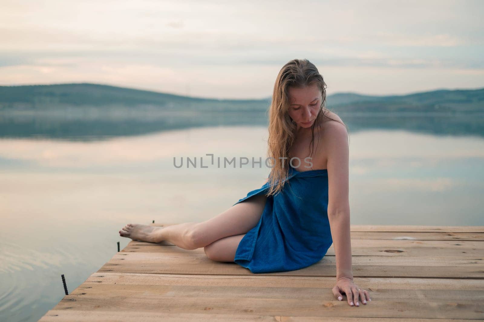 A young woman with wet hair, wrapped in a blue towel, sits on a wooden dock by a calm lake during sunset.