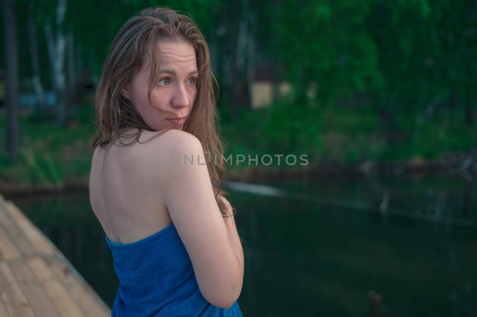 A young woman with wet hair wrapped in a blue towel stands near a calm lake at sunset, gazing thoughtfully into the distance.