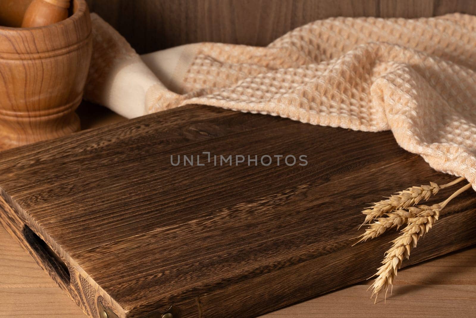 A dark wooden cutting board rests on a rustic table, accompanied by a woven towel and a bowl of wooden utensils, adding warmth to the kitchen setting with grains nearby.