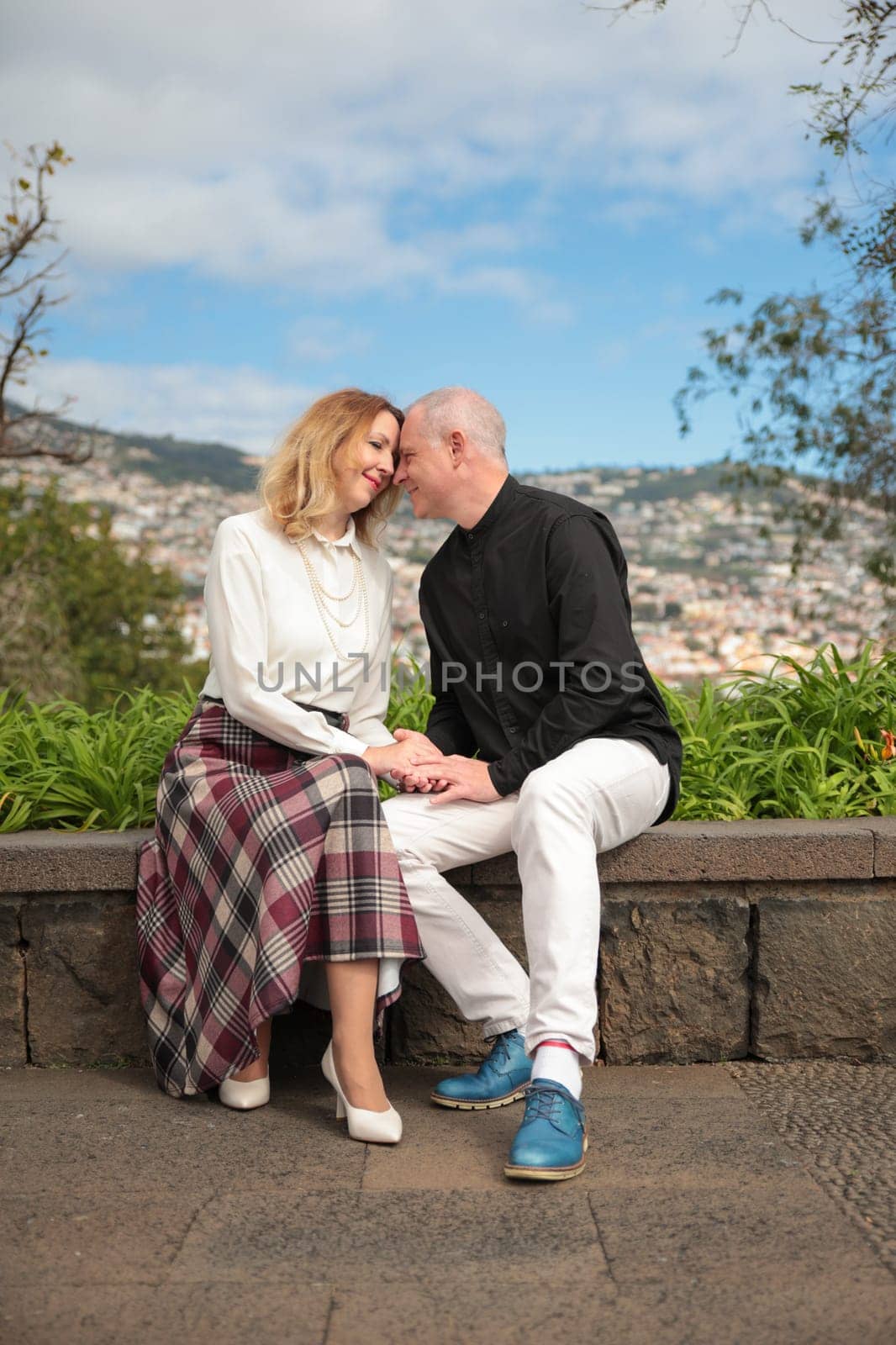 Happy mature couple holding hands and touching foreheads while sitting on a stone wall, enjoying the panoramic view of funchal
