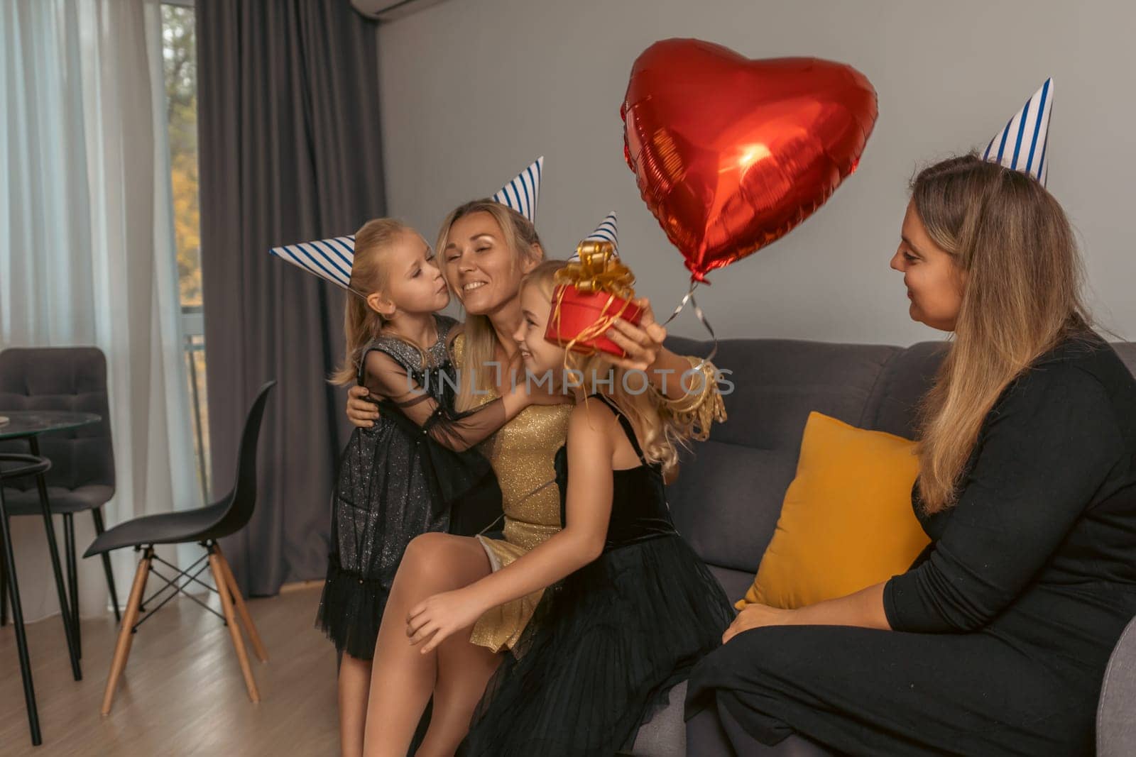 A woman in a gold dress receives kisses and a gift from two children wearing party hats, while another woman watches a red heart balloon floats nearby. Happy birthday celebration.