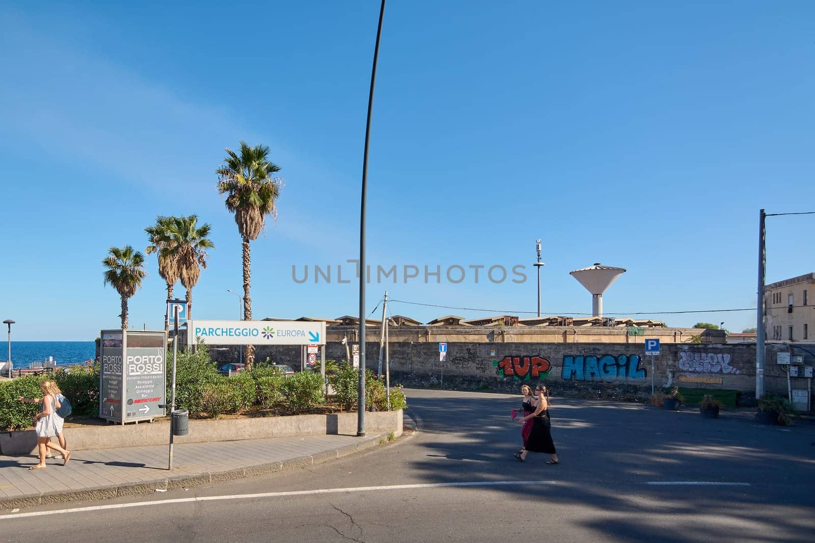 Catania. Italy - February 25, 2025: A sunny day at a coastal parking area in Catania with palm trees, graffiti on the walls, and people walking. Concept: Urban landscape