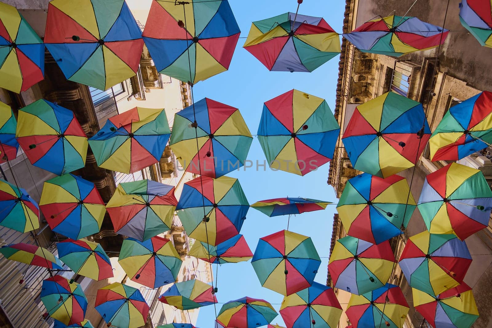 A dazzling array of colorful umbrellas hangs under a sunny sky, adding cheer to the urban scene. Concept: Playful sky decoration