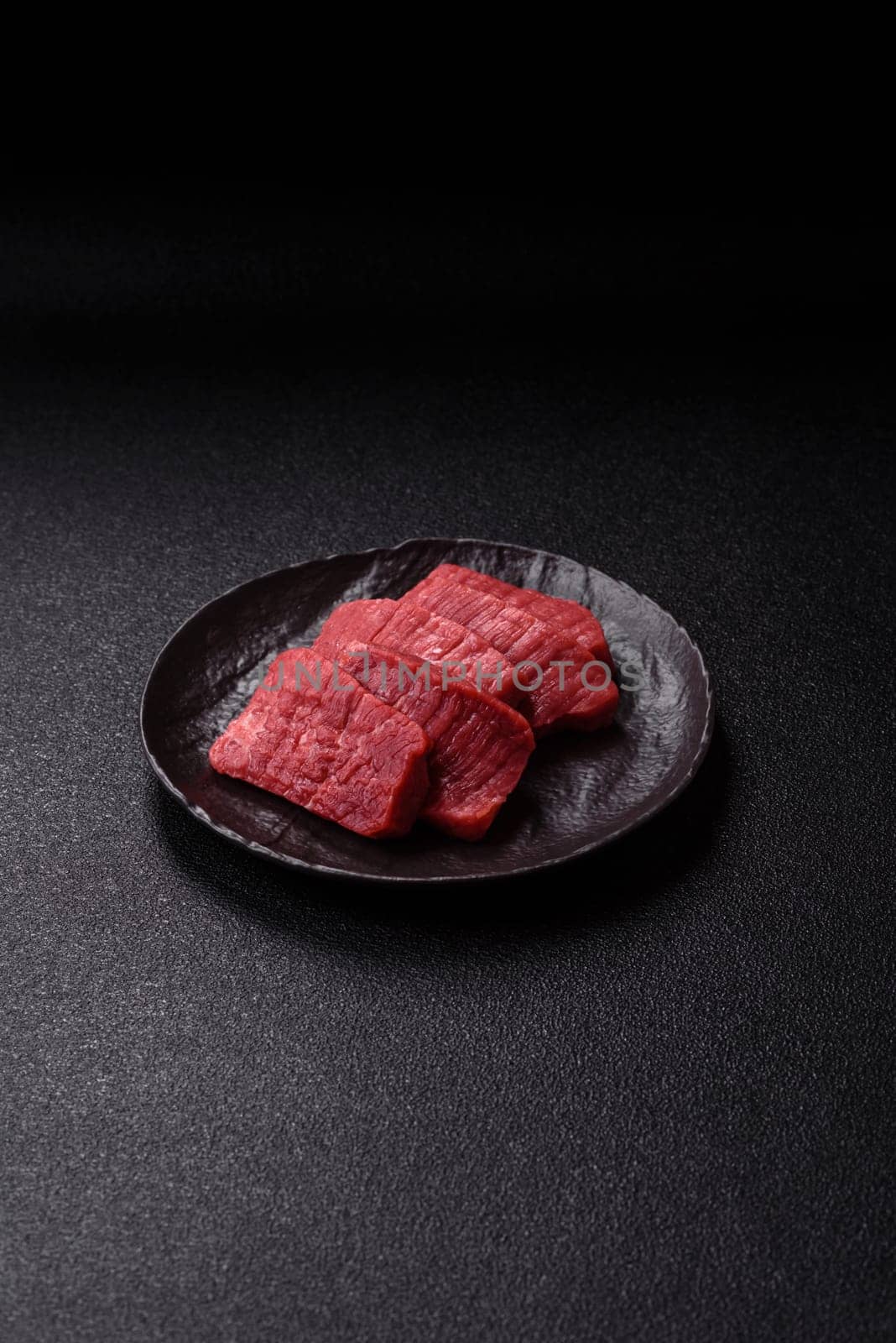 Raw beef tenderloin cut into slices with salt and spices on a dark concrete background