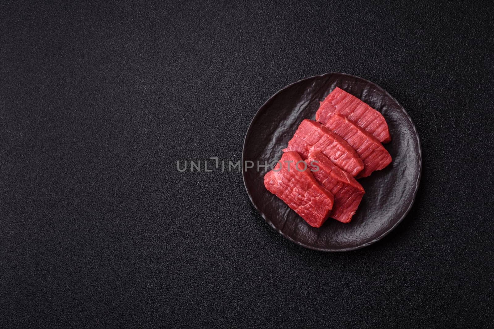Raw beef tenderloin cut into slices with salt and spices on a dark concrete background