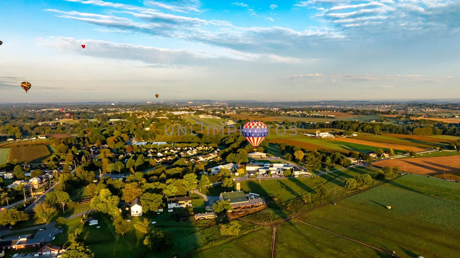 Hot air balloons of various colors drift gracefully over a scenic countryside at sunrise. The serene landscape features fields, trees, and quaint houses, creating a beautiful morning view.
