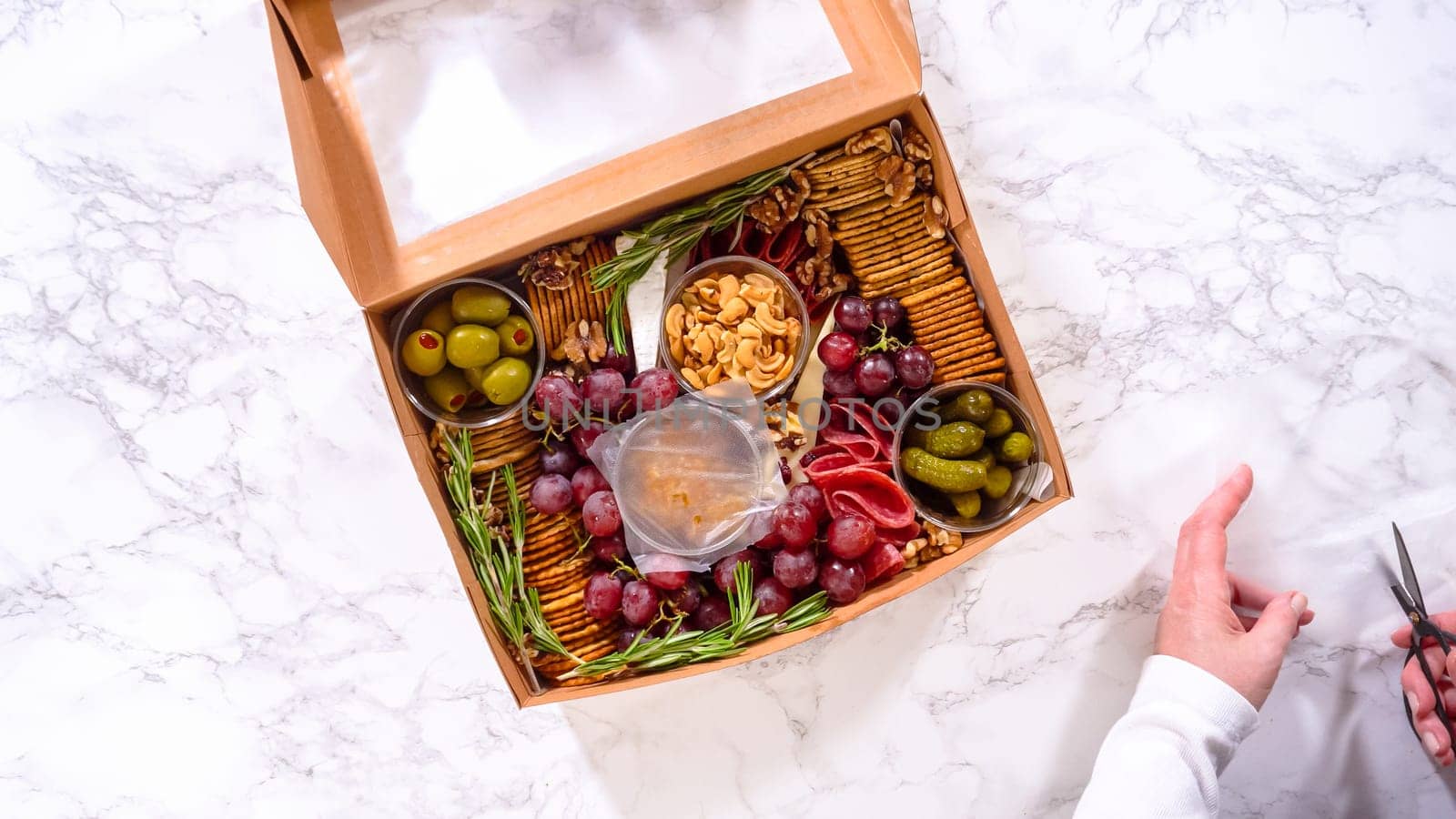 Flat lay. Hands are meticulously adding fresh red grapes to a bowl, complementing a beautifully arranged charcuterie box brimming with a variety of cheeses, olives, and cured meats, set against a sleek marble surface.