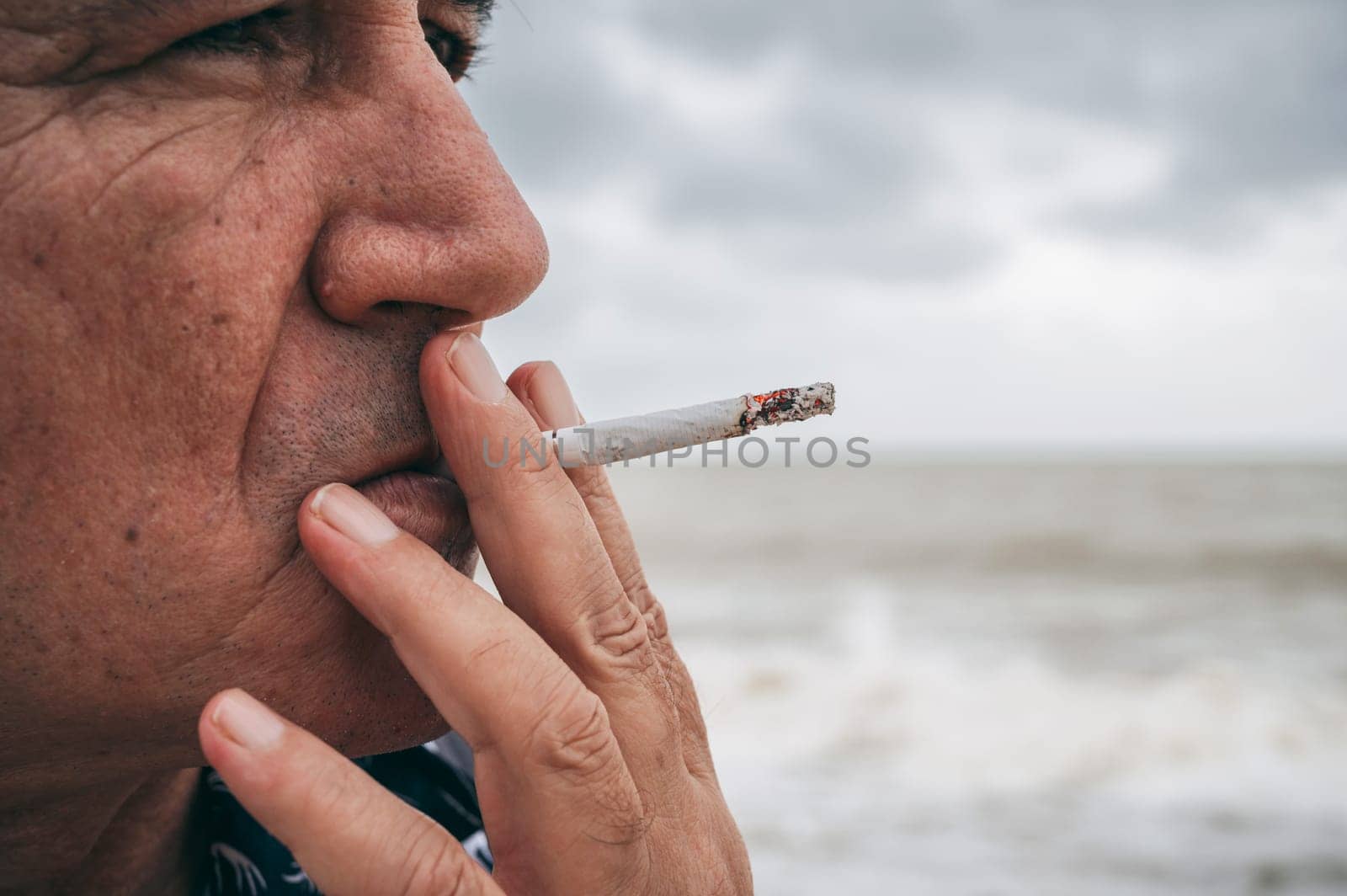portrait of a sad adult elderly Caucasian man smoking a cigarette alone outdoors on the beach close-up