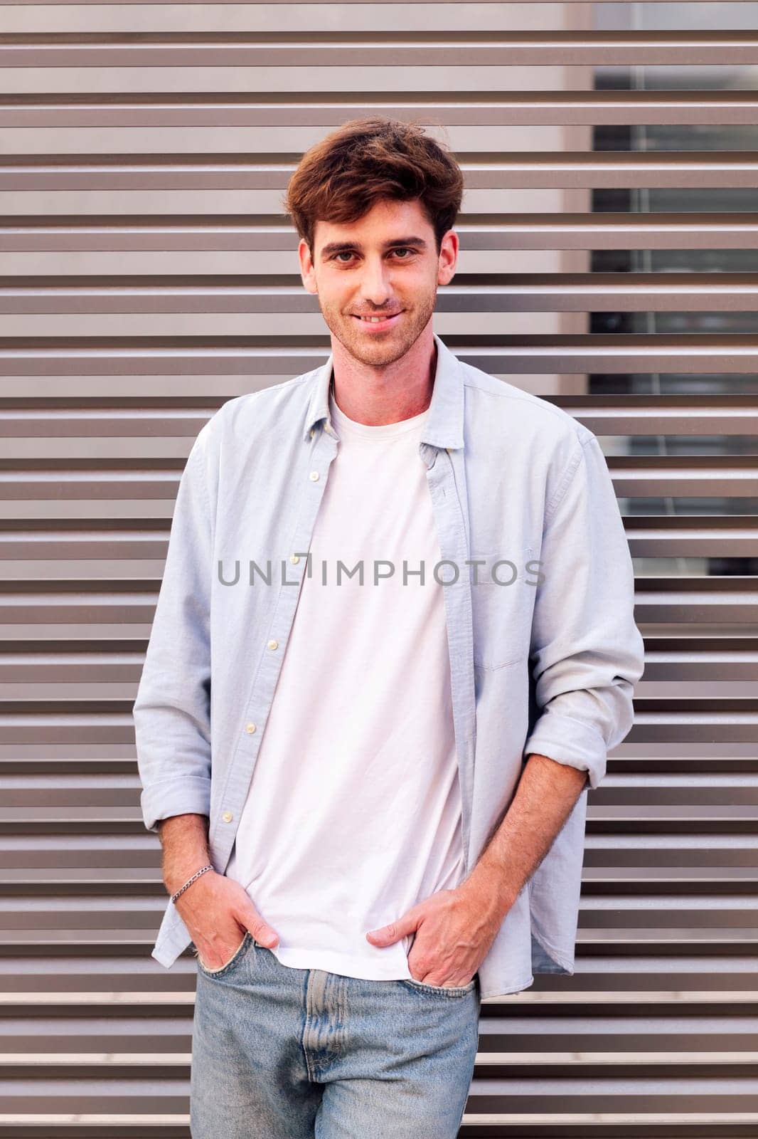 man in white shirt and blue jeans smiling looking at camera with hands in pockets, he is standing with a gray wall at background, concept of youth and urban lifestyle