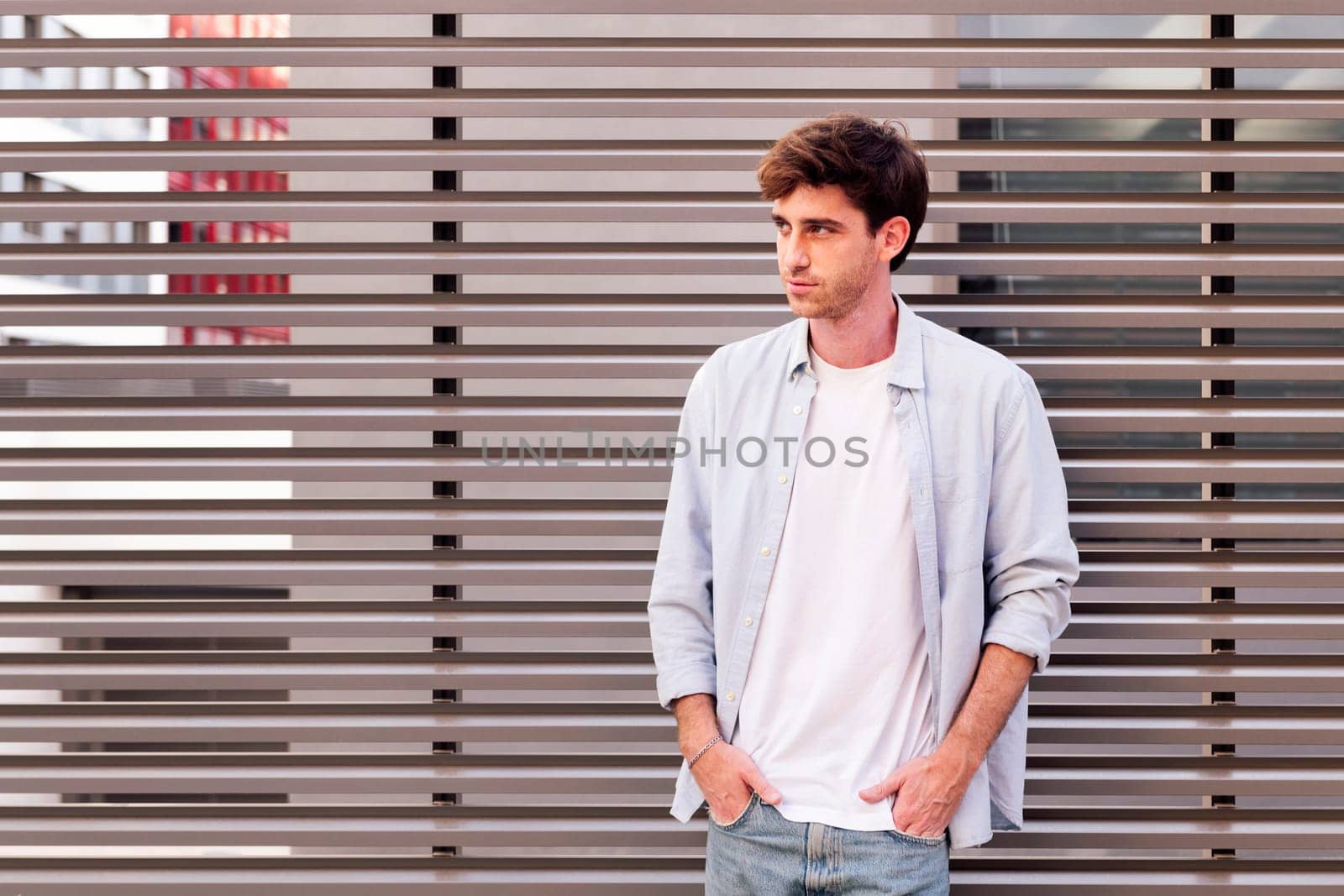 man in white shirt and blue jeans smiling looking away with hands in pockets, he is standing with a gray wall at background, concept of youth and urban lifestyle, copyspace for text