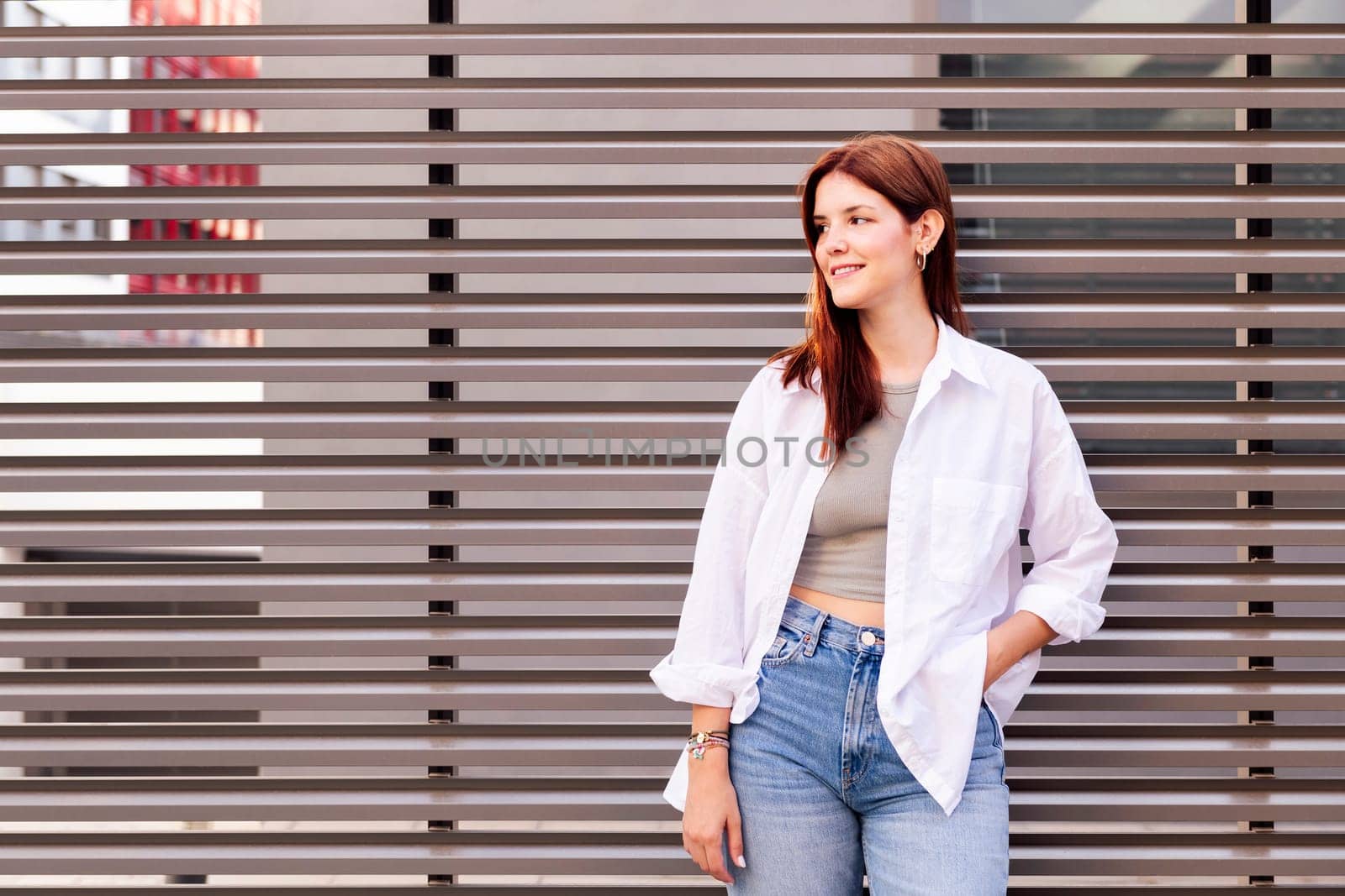 woman in white shirt and blue jeans smiling looking away with hands in pockets, she is standing with a gray wall at background, concept of youth and urban lifestyle, copyspace for text