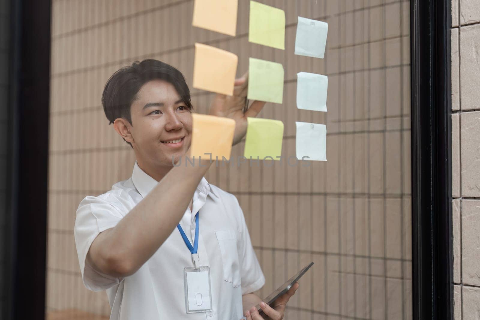 A young professional is smiling as he arranges colorful sticky notes on a glass wall, effectively organizing tasks and improving workflow in a modern office environment.