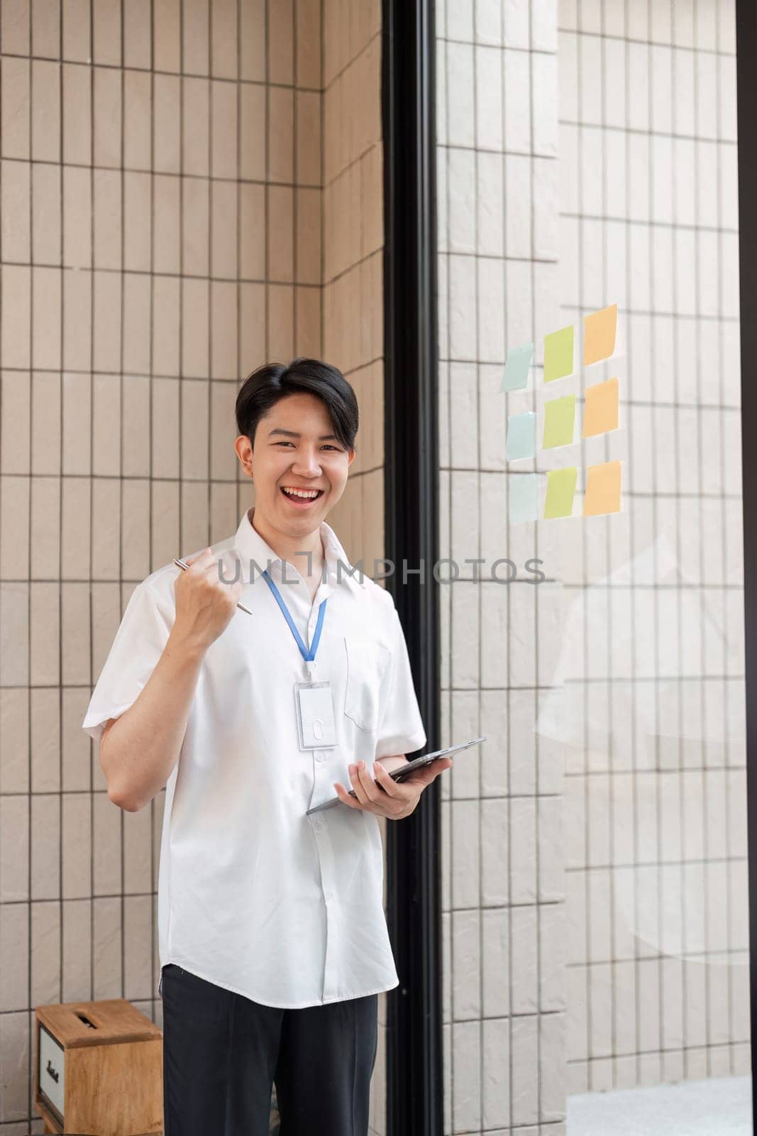 A young professional is joyfully celebrating success while holding a tablet, standing beside an organized array of colorful sticky notes on a glass wall in a contemporary office.