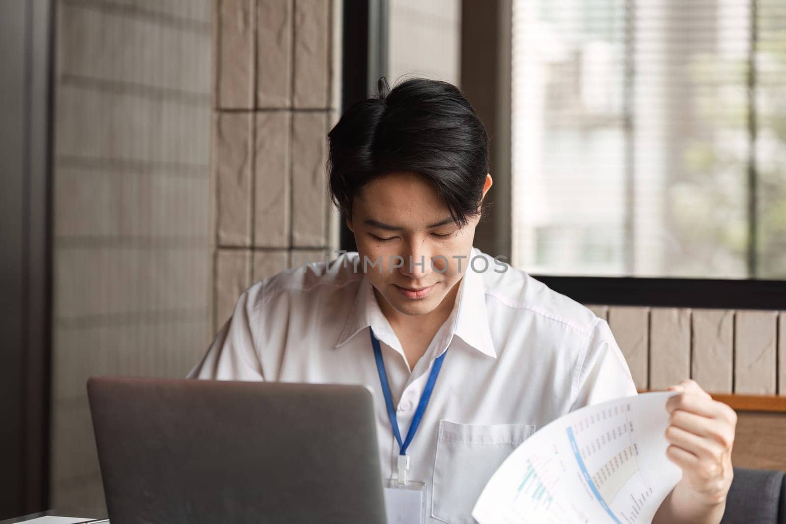 A young businessman is focused on analyzing financial reports while working on his laptop in a contemporary office space.
