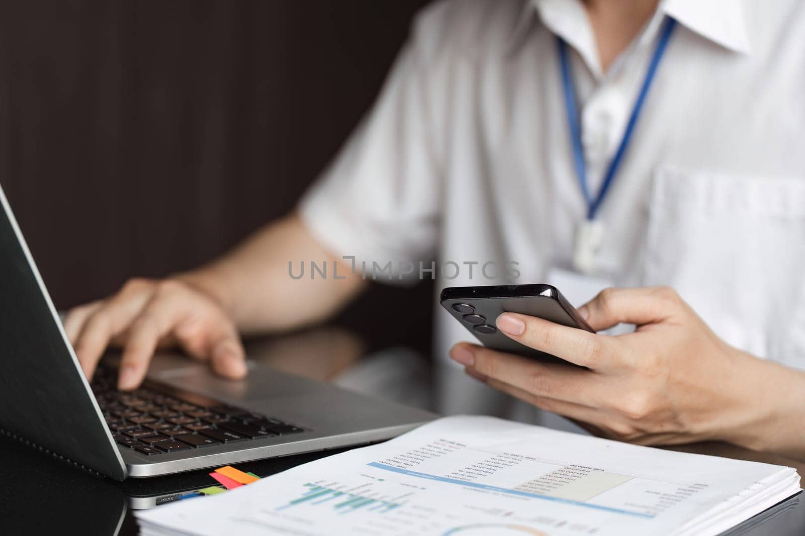 A young Asian accountant multitasks, using a smartphone in one hand while working on a laptop and reviewing financial documents in a modern office.