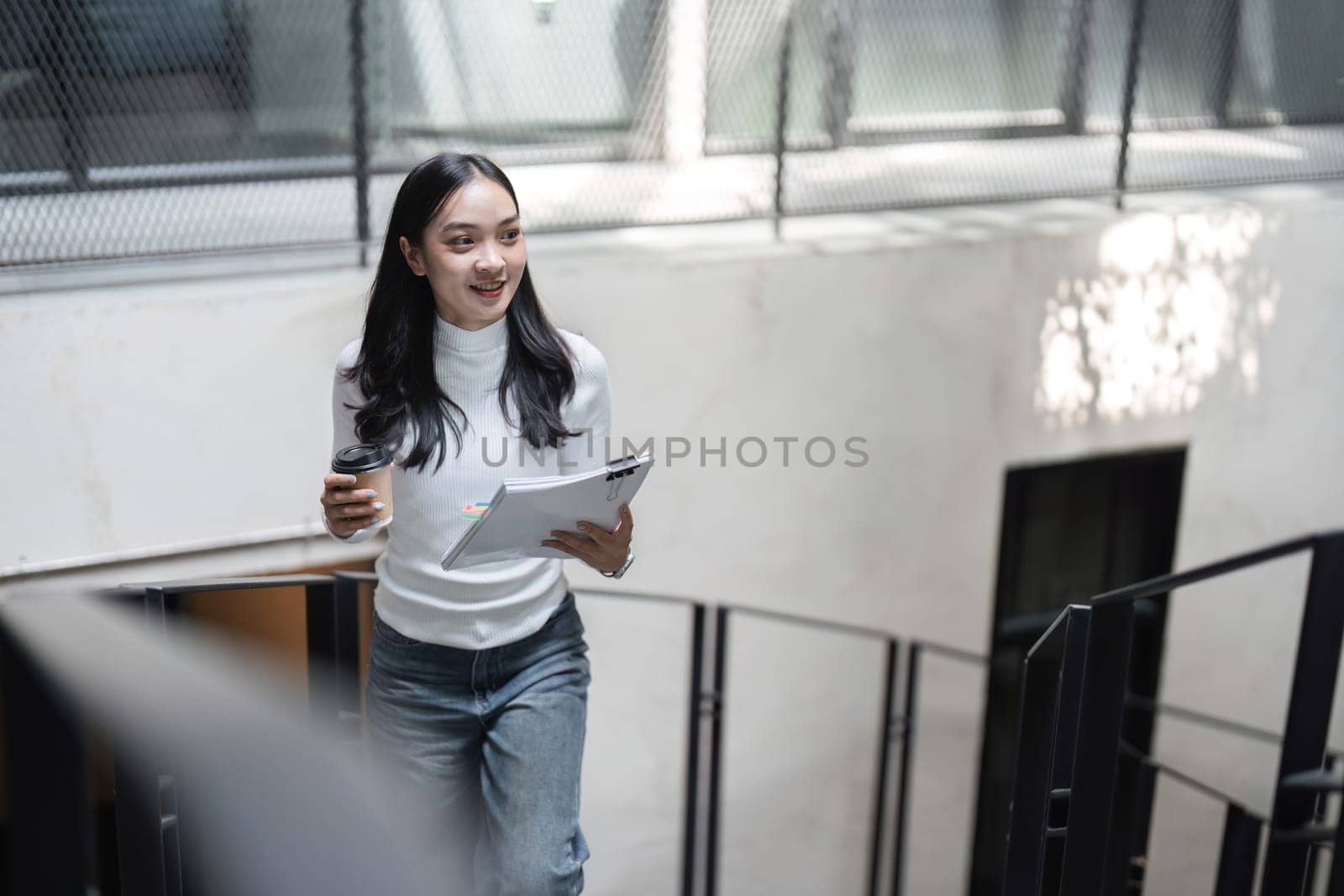 A young professional woman is walking up the stairs, holding a coffee cup and documents while smiling, embodying a positive and focused attitude in a contemporary office.