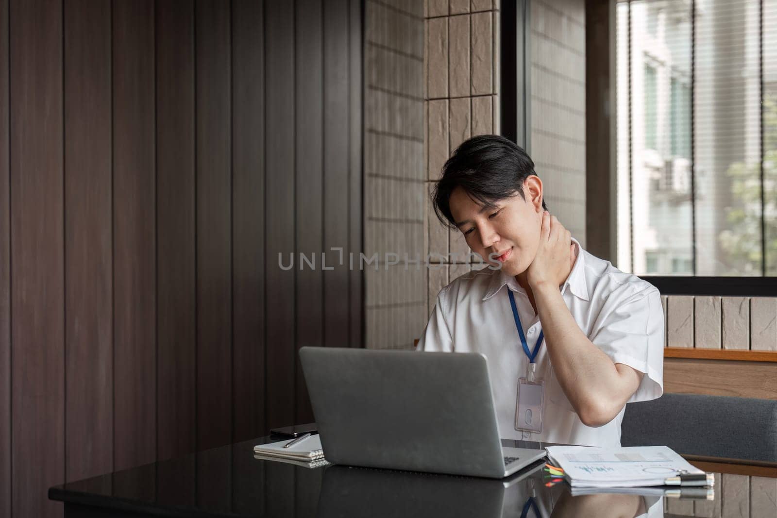 A young professional is rubbing his neck in discomfort while working on his laptop in a contemporary office, emphasizing the need for ergonomic practices.