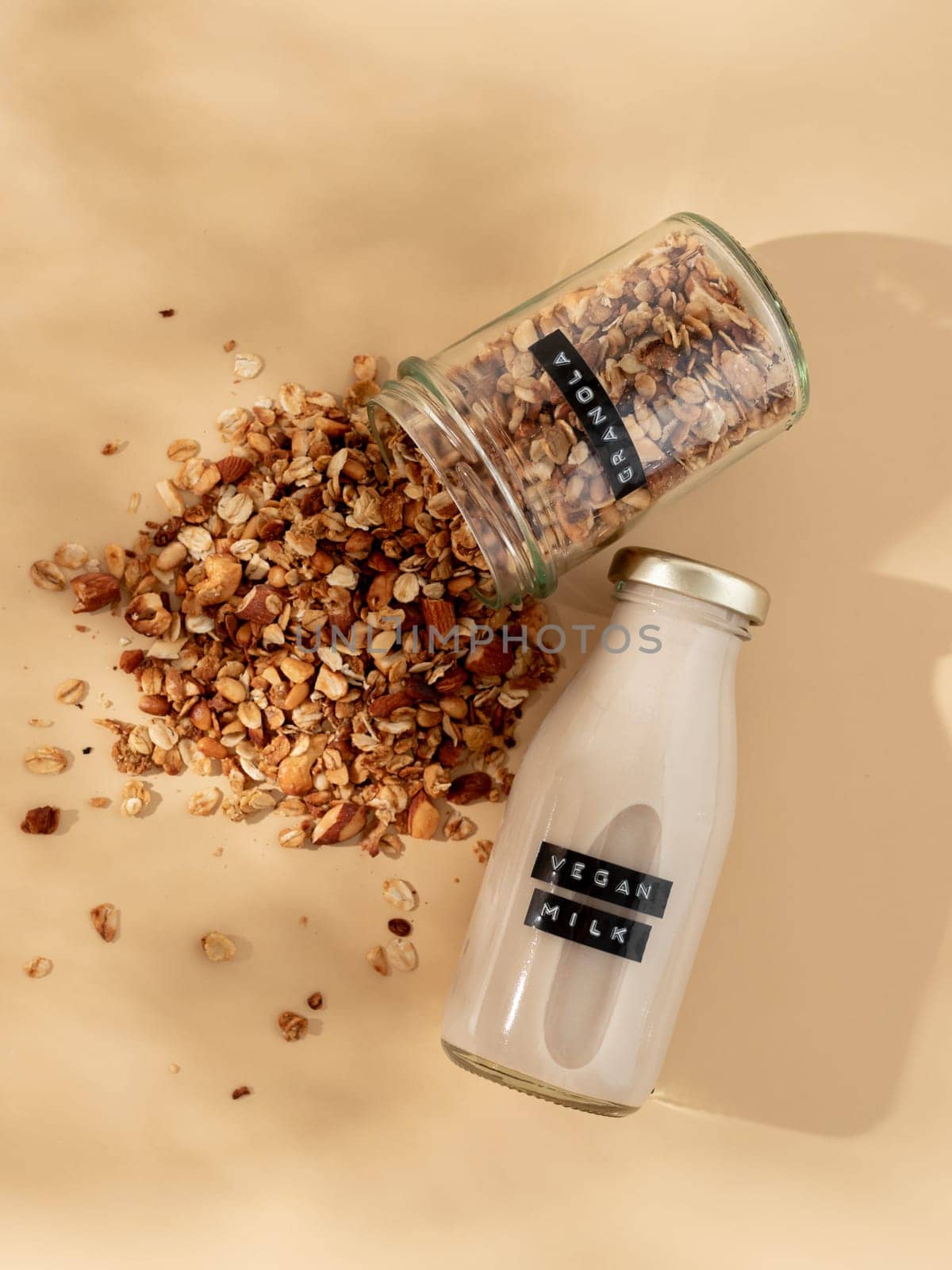 Granola and vegan milk. Healthy breakfast aesthetic concept. Homemade granola sprinkled of glass jar and vegan milk in glass bottle over champagne yellow or pastel beige background. Top view flat lay