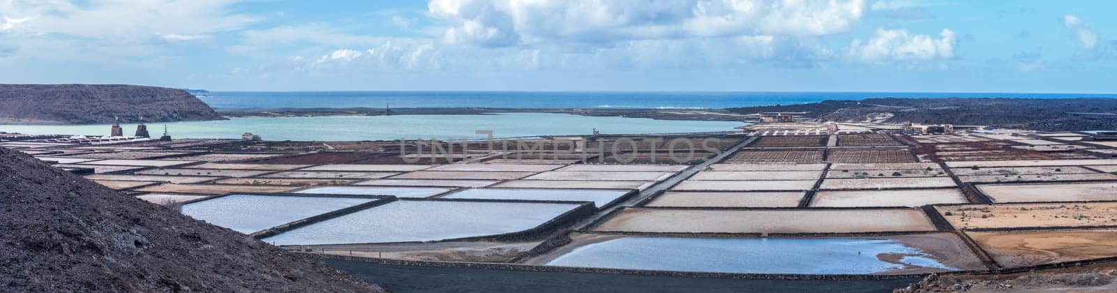 Vast salt flats extend towards the ocean under a cloudy sky, forming an intriguing and textured landscape.