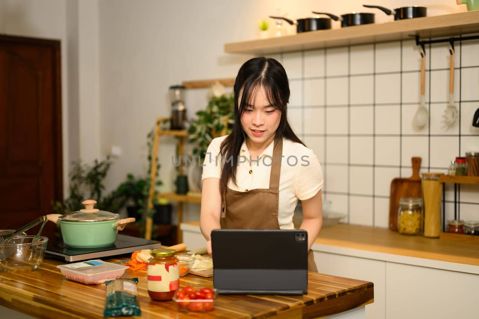 Young Asian woman following a spaghetti recipe on digital tablet while cooking in cozy kitchen.