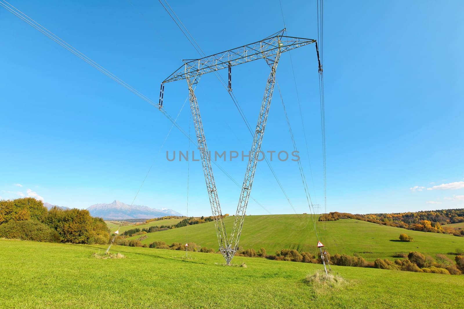 Large metal power pylon under electricity lines built in countryside with green grass covered hills and small forests, mount Krivan (Slovak symbol) at distance, blue sky background 