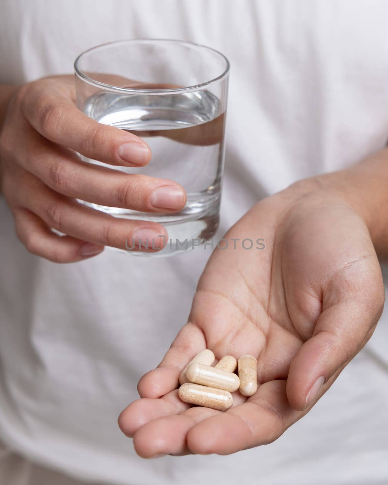 Woman in white t-shirt holding beige capsules in cupped hand and water glass in hand closeup. Female or male taking dietary supplements or vitamins. Healthcare concept