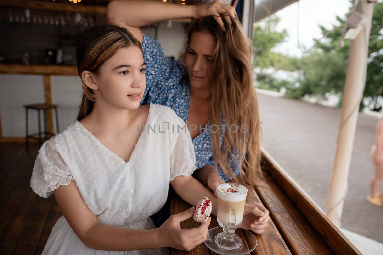 Two women sit at a cafe. One woman's hair is being adjusted by the other. They share a latte. A relaxed, intimate moment.