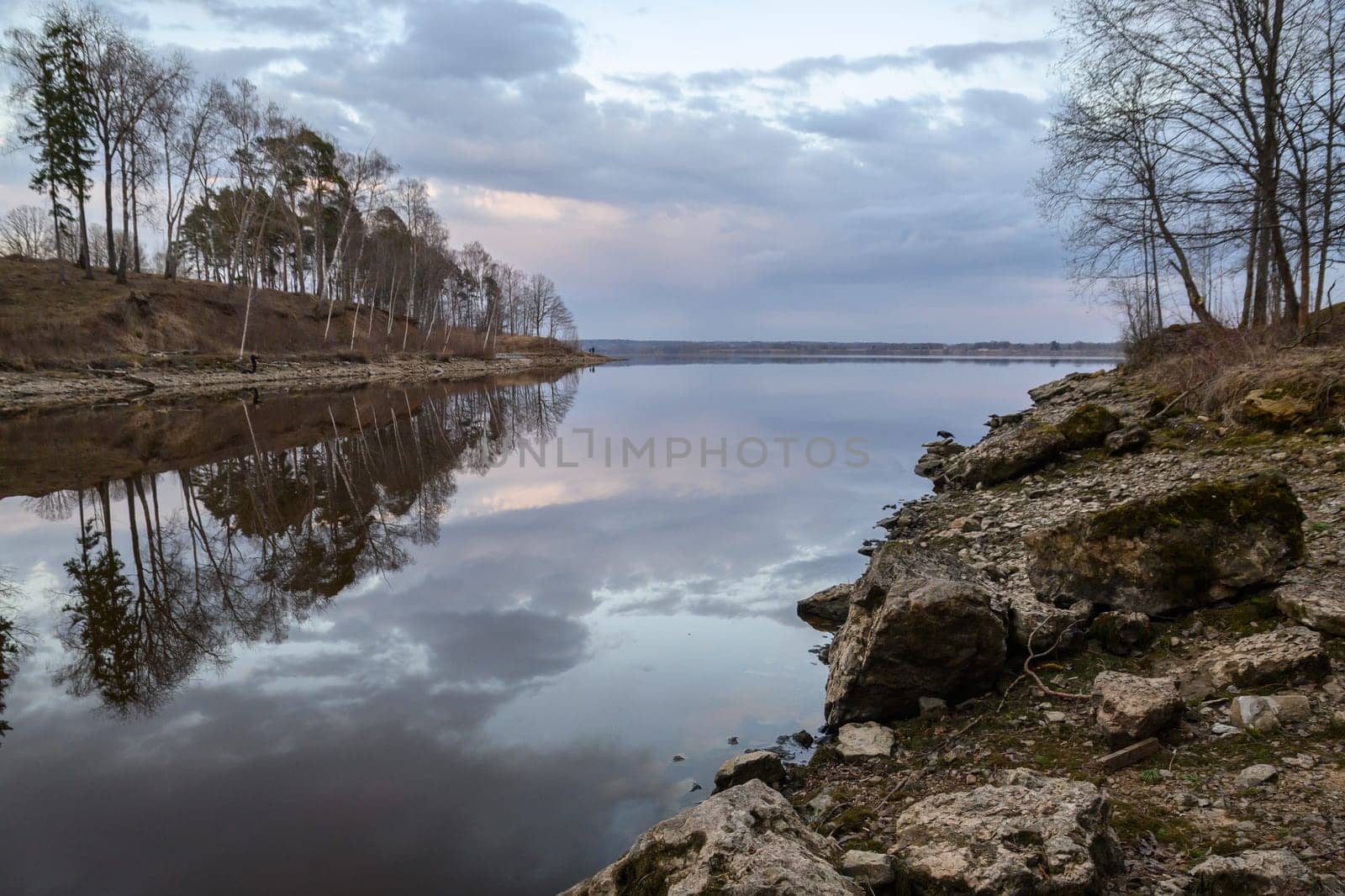 Soft hues of evening light dance across still waters, framed by trees and rugged stones along the riverbank.