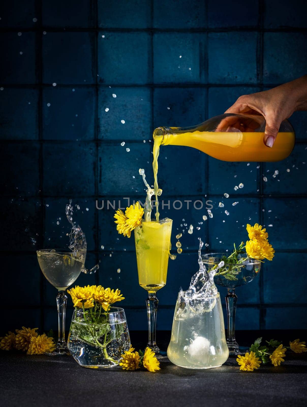 Bartender pours a bright yellow cocktail into various glasses, creating splashes and spills, adorned with yellow flowers against a blue tile backdrop