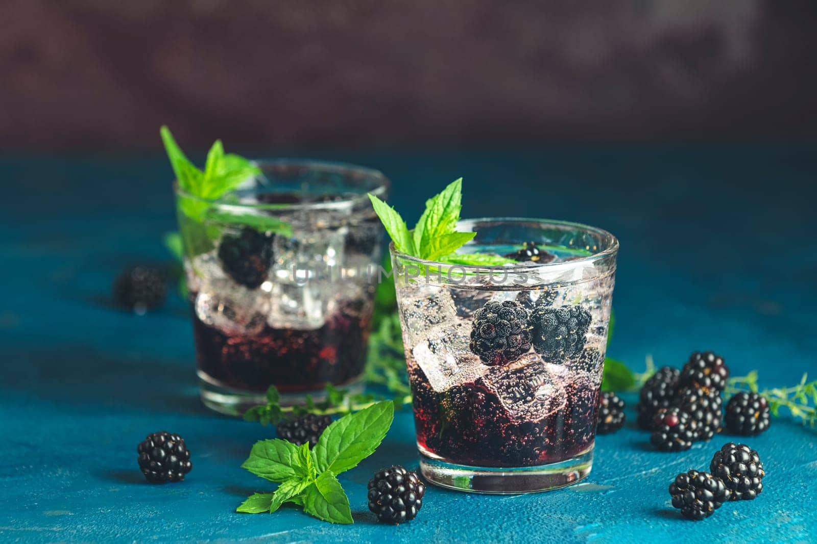 Two glasses of sparkling blackberry cocktail with ice cubes, fresh mint and blackberries on blue background
