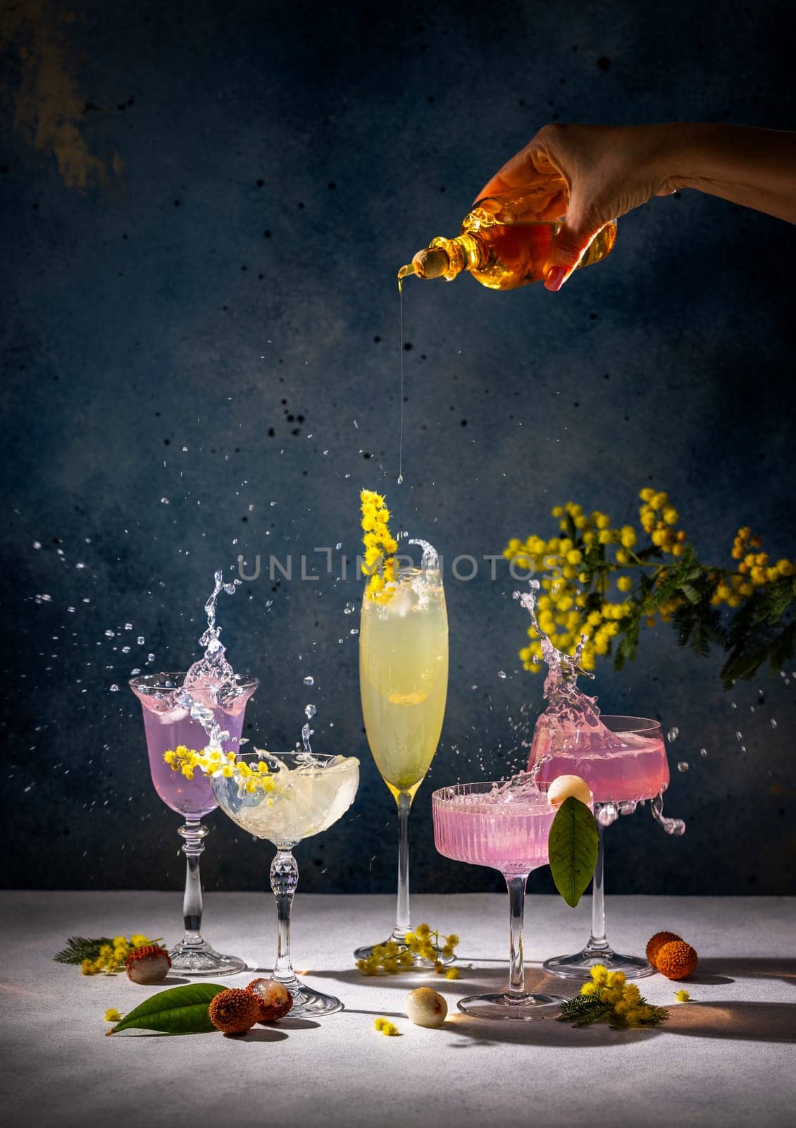Syrup being poured into various cocktails, creating splashes and adorned with mimosa flowers, lychees, and a textured backdrop