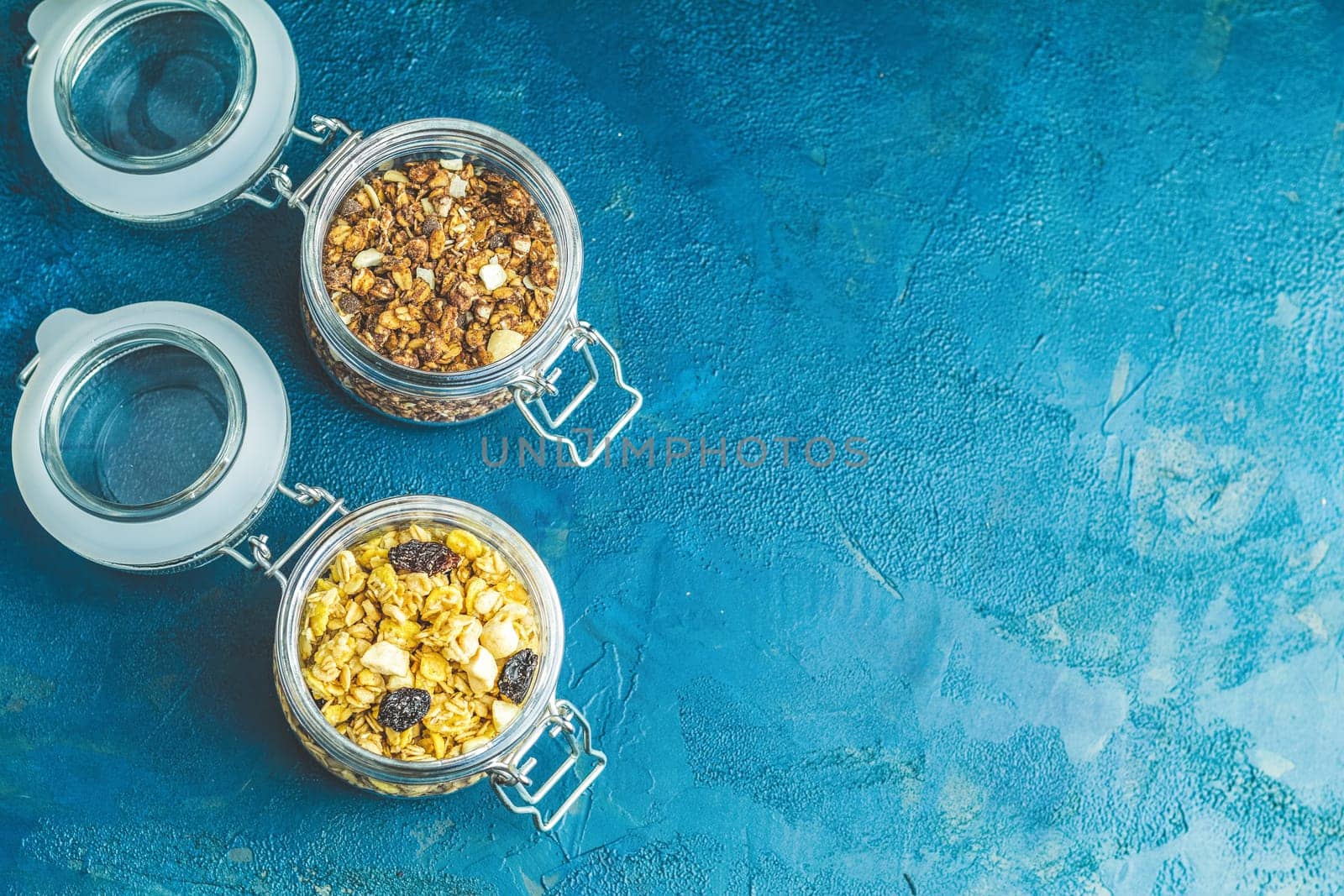 Two open glass jars containing chocolate and classic homemade granola on rustic blue background with copy space