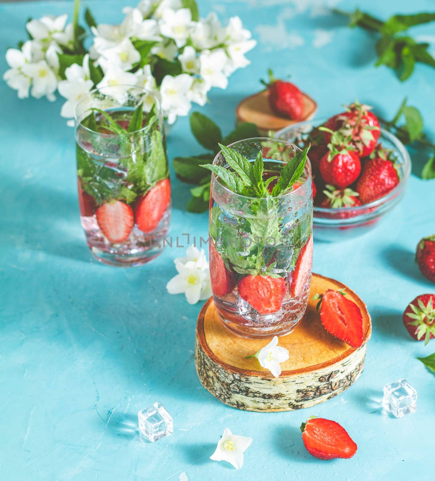 Two glasses of refreshing infused water with fresh strawberries and mint on a light blue background, decorated with jasmine flowers