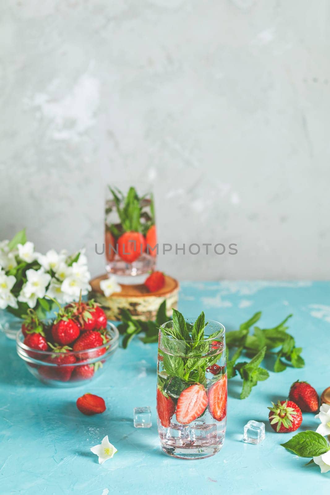 Two glasses of refreshing infused water with ripe strawberries and mint leaves are standing on a light blue background