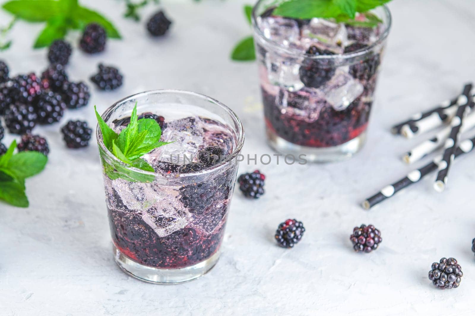 Two glasses of refreshing blackberry cocktail with ice, mint and black and white straws on a light background