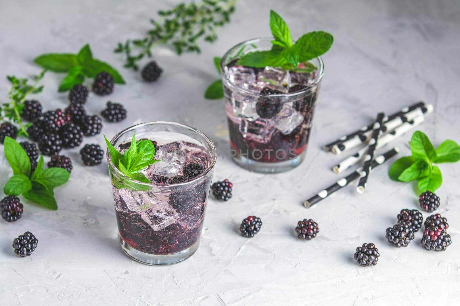 Two glasses of blackberry cocktail, garnished with mint and ice, sit among fresh blackberries, mint sprigs, and thyme on a textured surface