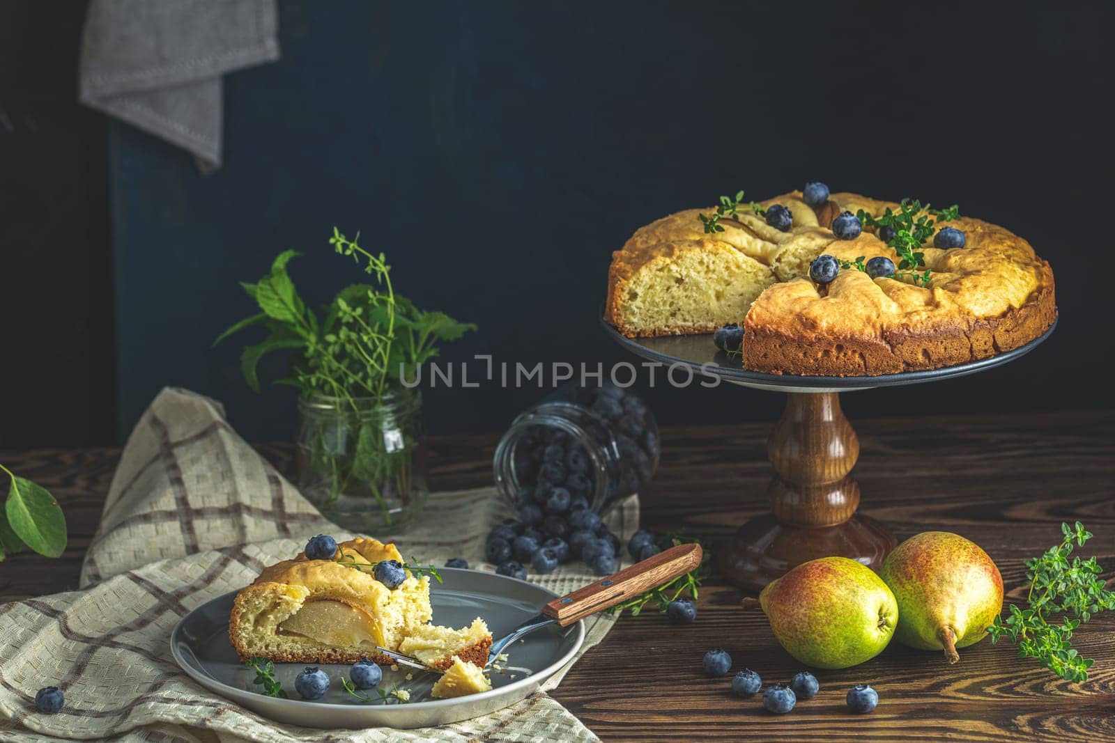 Homemade pear and blueberry cake decorated with fresh thyme and blueberries is served on a rustic wooden table