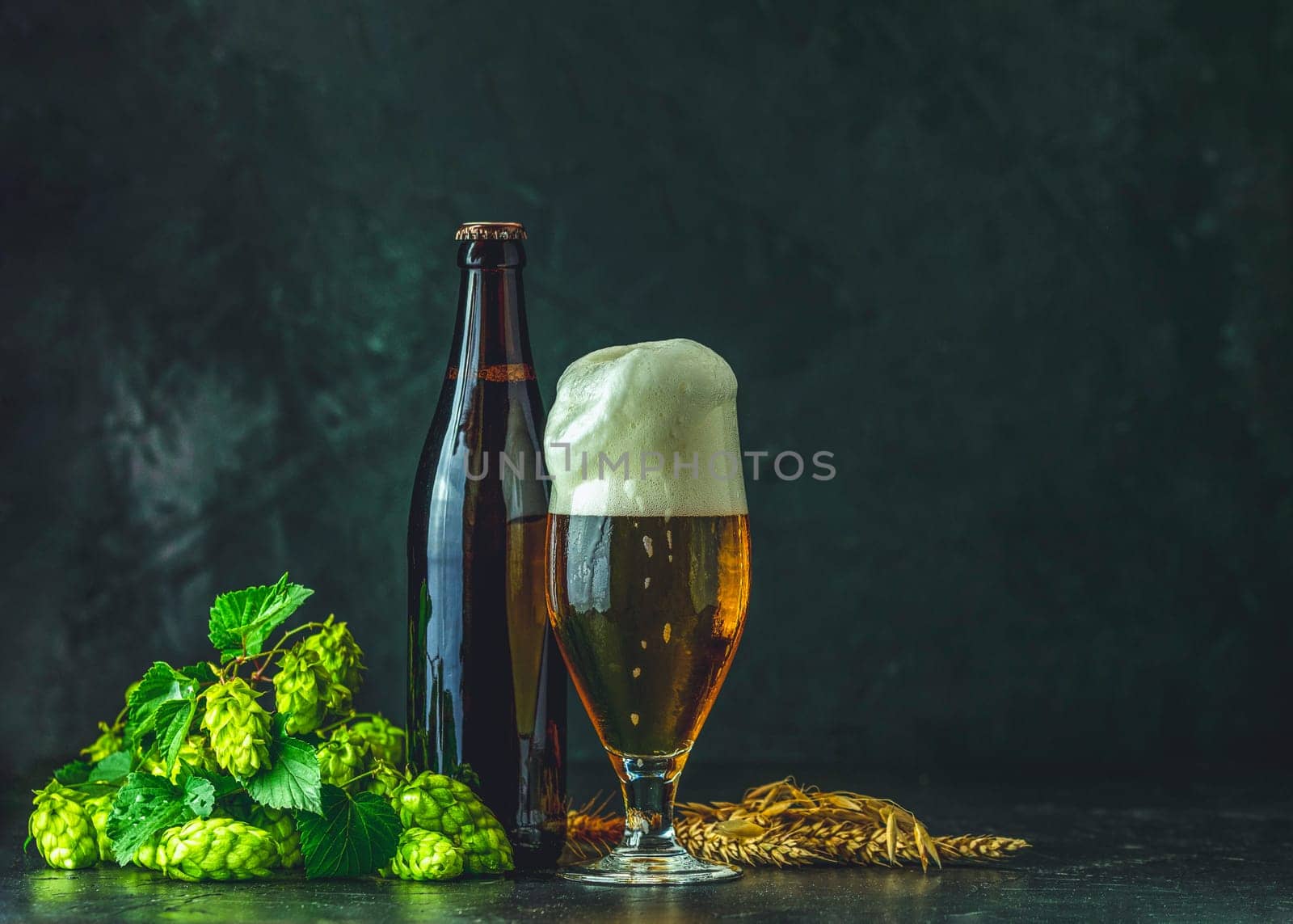 Beer glass and dark bottle with overflowing foam, fresh hops and wheat ears on dark background representing brewing process