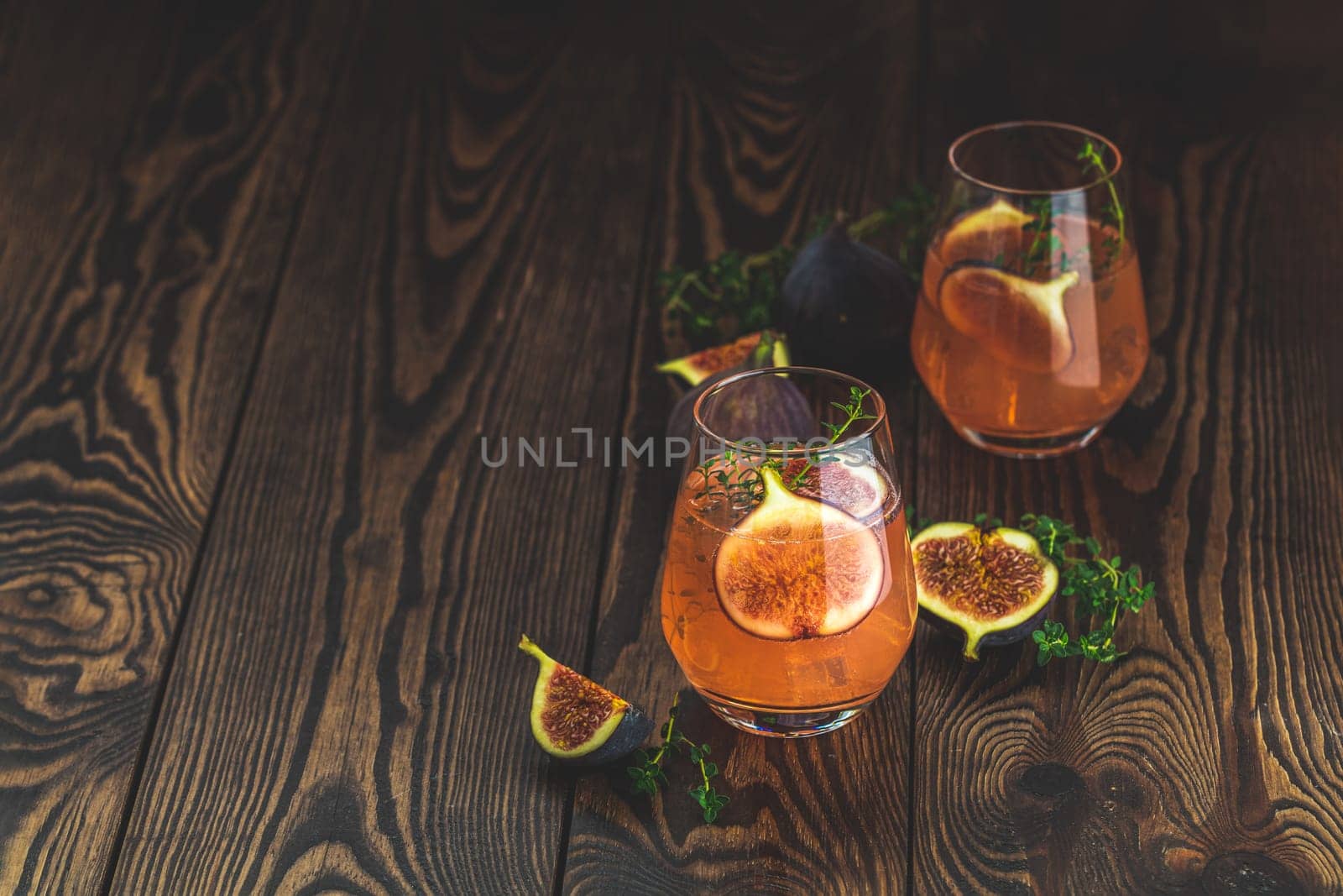 Two refreshing pink cocktails garnished with fresh figs and thyme sprigs sit on a rustic wooden table, creating a vibrant and inviting scene