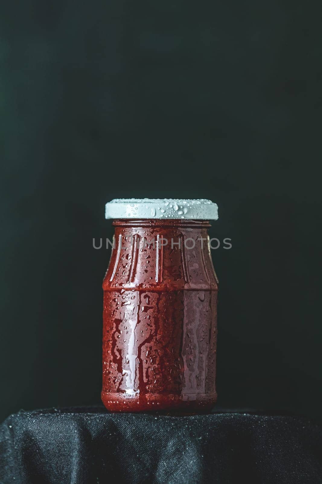 Small glass jar containing red sauce, covered with condensation, sits on a dark cloth against a black background