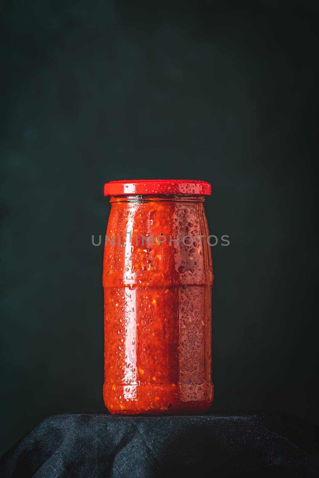 Glass jar full of traditional balkan vegetable spread ajvar or pindjur, with drops of condensation on glass, on black background