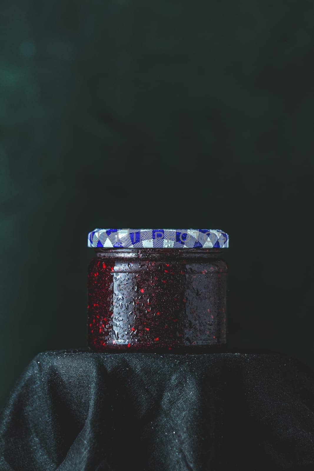 Glass jar of red raspberry jam with condensation and checkered lid sits on black cloth against a dark background