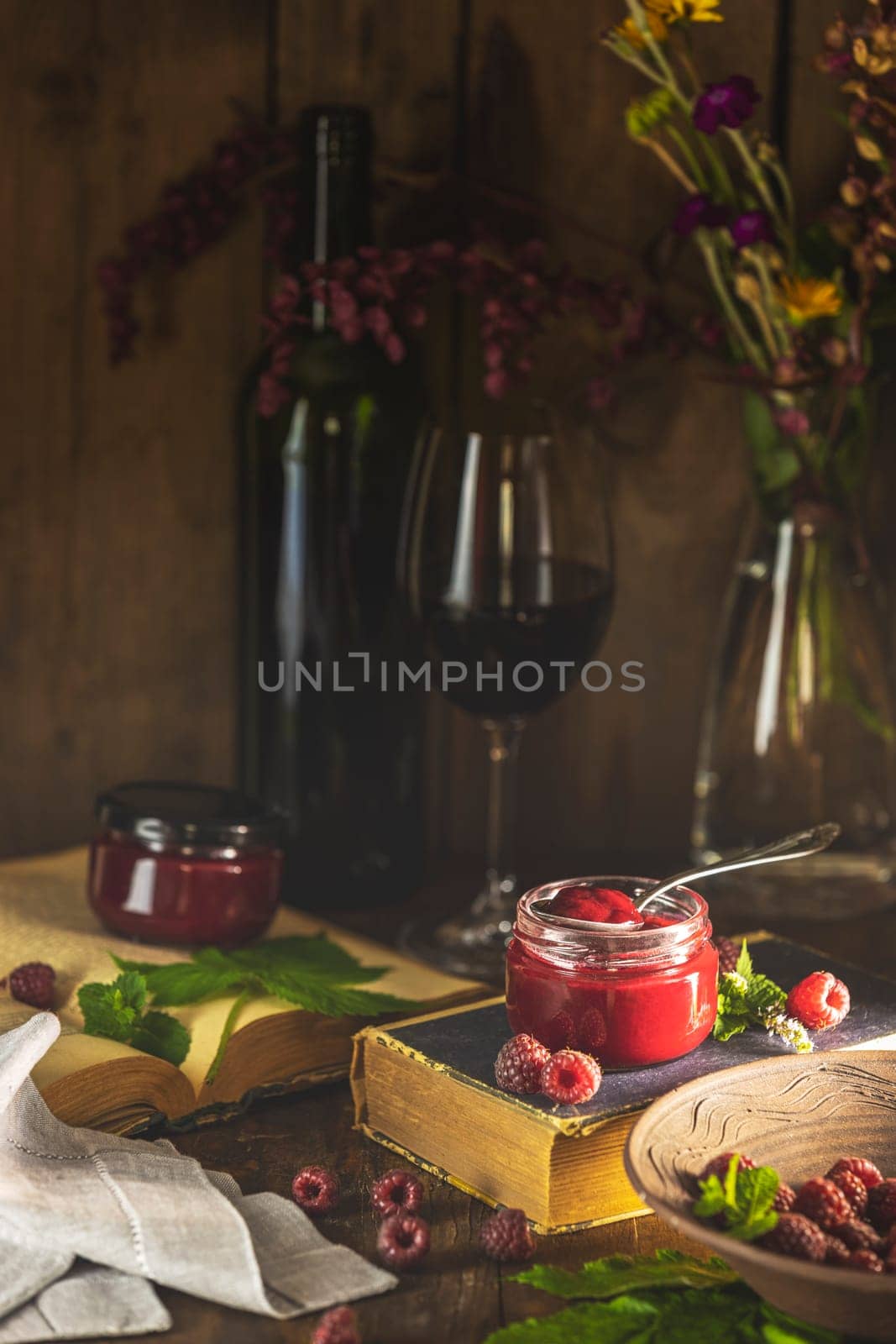 Homemade raspberry jam in a glass jar with fresh raspberries, mint leaves, a bottle of wine, a glass of wine, old books, and flowers on a wooden table