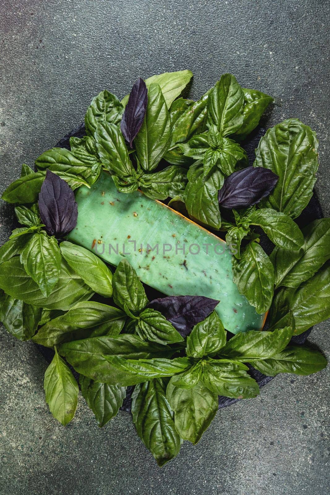 Green pesto gouda cheese decorated with fresh green and purple basil leaves on dark background, top view
