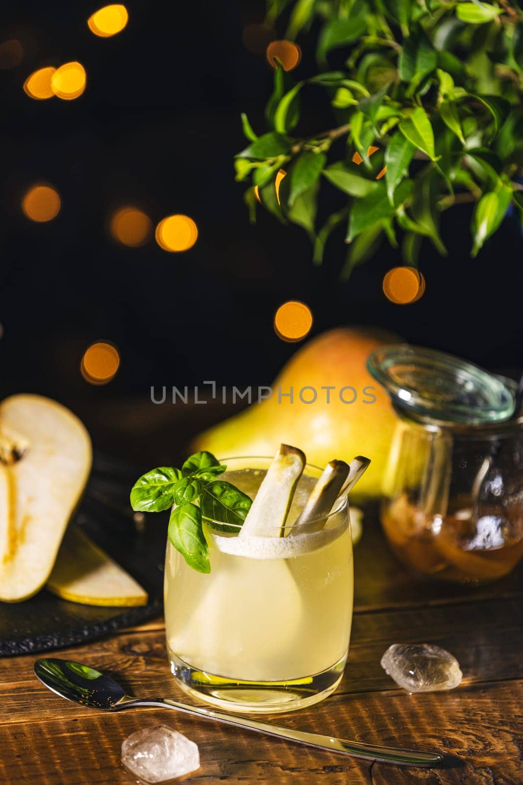 Pear cocktail with basil and fruit slices is served on a wooden table with ice cubes, a spoon, and a jar of pear jam in the background with bokeh effect
